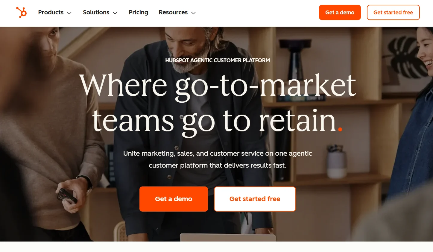HubSpot homepage showcasing its AI-powered customer platform with the headline "Where go-to-market teams go to retain," featuring professionals collaborating around a laptop and prominent "Get a demo" and "Get started free" call-to-action buttons.